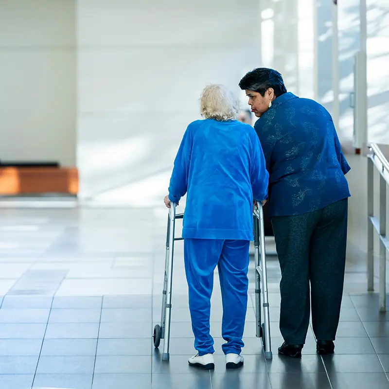 An older woman with a walker being escorted down a well lit hallway